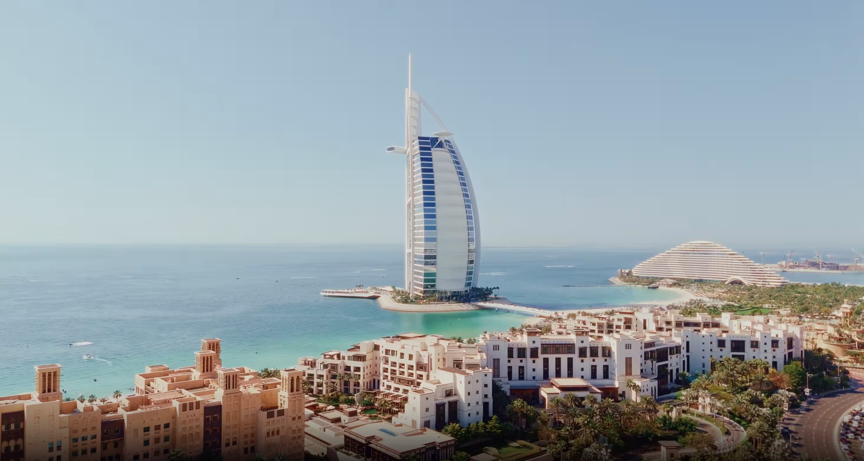 Aerial view of the Burj Al Arab hotel surrounded by turquoise sea and nearby coastal buildings under a clear blue sky.